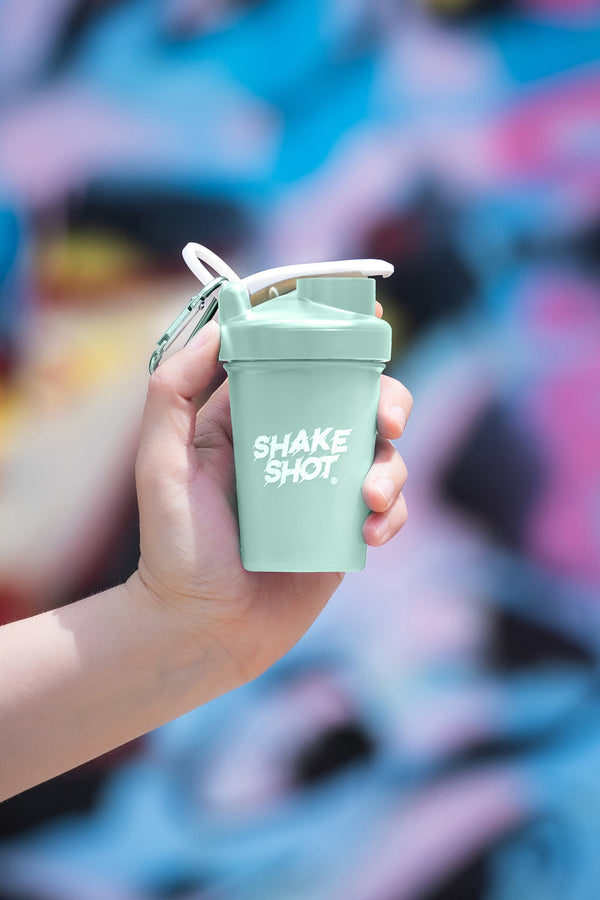 A green mini shaker bottle labeled 'Shake Shot' being held by a hand, with a carabiner attached to it, against a colorful background.