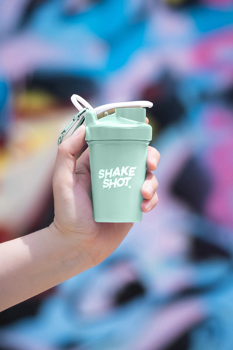 A green mini shaker bottle labeled 'Shake Shot' being held by a hand, with a carabiner attached to it, against a colorful background.
